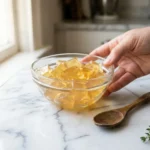 Glass bowl of golden gelatin on white marble surface with wooden spoon