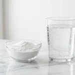 baking soda in a bowl next to a glass of water on white marble