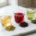 Three glass cups of chamomile, hibiscus, and peppermint herbal tea with dried herbs on white marble