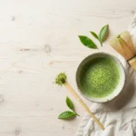 Bowl of frothy green matcha tea with a bamboo whisk and scoop of matcha powder