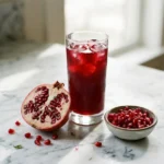 Tall glass of deep red pomegranate juice with ice beside a halved pomegranate and a bowl of arils on white marble