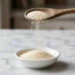 Wooden spoon lifting unflavored gelatin powder over a small white bowl on white marble